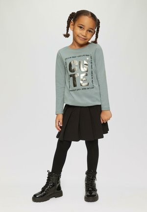 Lange mouw teal shirt met metallic "CUTE" tekst, gecombineerd met een zwarte geplooide rok, zwarte panty en glanzende zwarte veterschoenen.