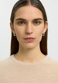 Joven mujer con el pelo castaño liso partido por la mitad, lleva pendientes pequeños de aro plateados y una camiseta beige de cuello redondo, mirando hacia adelante.