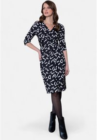 Robe noire enveloppante ornée de motifs abstraits blancs, à manches trois-quarts, avec une taille à nouer et un ourlet au genou. Portée avec des bottines.