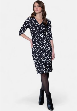 Robe noire enveloppante ornée de motifs abstraits blancs, à manches trois-quarts, avec une taille à nouer et un ourlet au genou. Portée avec des bottines.
