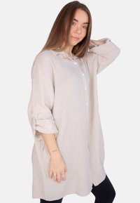 Hellbeige Langarmhemd aus strukturiertem Stoff, mit Knopfleiste und hochgekrempelten Ärmeln, kombiniert mit dunklen Hosen.