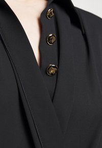 Blusa nera con scollo a V, caratterizzata da quattro bottoni dorati con elementi decorativi e un tessuto liscio e testurizzato.