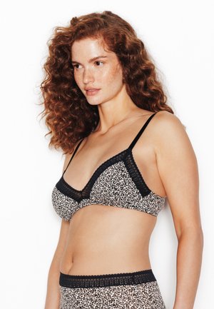 Donna con capelli rossi ricci che indossa lingerie con stampa leopardata e bordo in pizzo nero, in piedi contro uno sfondo bianco semplice, guarda a sinistra.