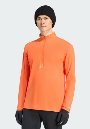 Uomo che indossa una maglia arancione brillante con zip e maniche lunghe, berretto nero, guanti neri e pantaloni neri su uno sfondo chiaro e uniforme.