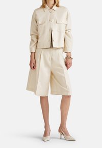 Beige zkrácená bunda se dvěma předními kapsami, kombinovaná s odpovídajícími širokými šortkami. Texturovaná tkanina, vyvážená silueta a minimalistický design.