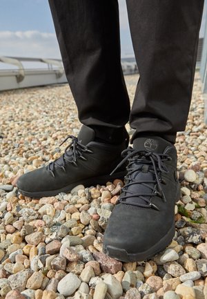 Osoba nosząca czarne buty trekkingowe Timberland stoi na żwirowej powierzchni na zewnątrz w czarnych spodniach.