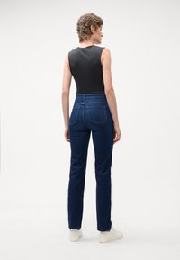 Temne modre kavbojke z visokim pasom in ravnim krojem, z zadnjimi žepi in gladkim, strukturiranim materialom. Model nosi črn top brez rokavov.