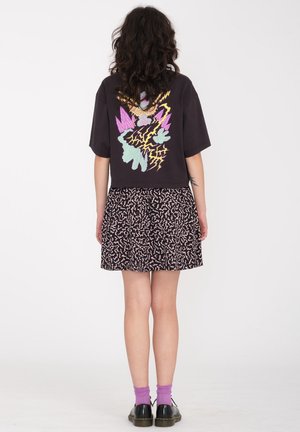 Femme aux cheveux bouclés portant un t-shirt noir avec un graphisme coloré, une jupe à motifs, des chaussettes violettes et des chaussures noires, debout de dos.