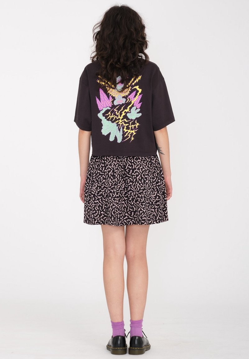 Femme aux cheveux bouclés portant un t-shirt noir avec un graphisme coloré, une jupe à motifs, des chaussettes violettes et des chaussures noires, debout de dos.