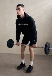 Haut à capuche noir avec le logo "Champion", shorts noirs et chaussures de sport noires. Le modèle soulève une barre avec des poids ronds. Fond neutre.