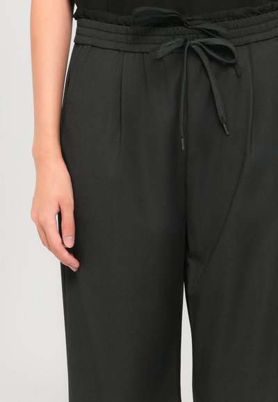 Left hand beside black drawstring pants with elastic waistband on a person against a light background.