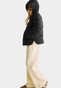 Giacca a vento nera con cappuccio, con un design compatto, texture trapuntata e polsini elasticizzati. Indossata sopra un pantalone chiaro.