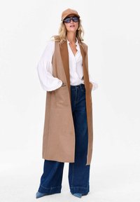 Długi brązowy kamizelka z dwoma przednimi kieszeniami, noszona na białej koszuli z bufkami, w połączeniu z szerokimi niebieskimi dżinsami i beżową czapką baseballową.