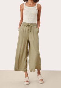 Szerokie spodnie w kolorze jasnej oliwki z elastycznym ściągaczem w talii i sznurkiem, zestawione z kremowym dzianinowym topem na ramiączkach. Materiał wydaje się miękki i lejący.