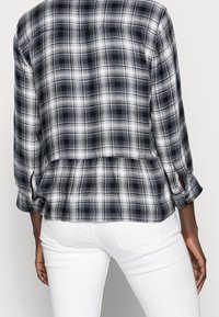 Chemise à carreaux noir et blanc avec manches retroussables et pli dans le dos. Fabriquée en tissu à la texture douce. Présente un ourlet fendu.