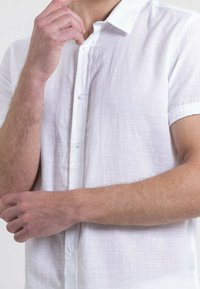 Chemise blanche à manches courtes, boutonnée, en tissu texturé et léger, avec un col classique et des boutons en plastique transparent.