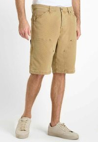 Shorts cargo beiges en tissu résistant, avec plusieurs poches, une coupe relaxée et des coutures visibles. Associés à des baskets beiges.
