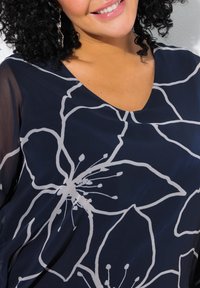 Navy blouse featuring large white floral patterns, a v-neckline, and sheer sleeves, made from lightweight fabric with a soft texture.