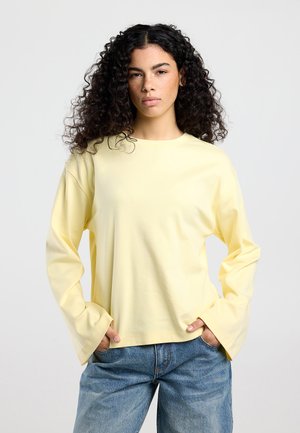 Jonge vrouw met krullend haar draagt een lichtgeel shirt met lange mouwen en blauwe spijkerbroek, staand met handen in de zakken tegen een effen achtergrond.