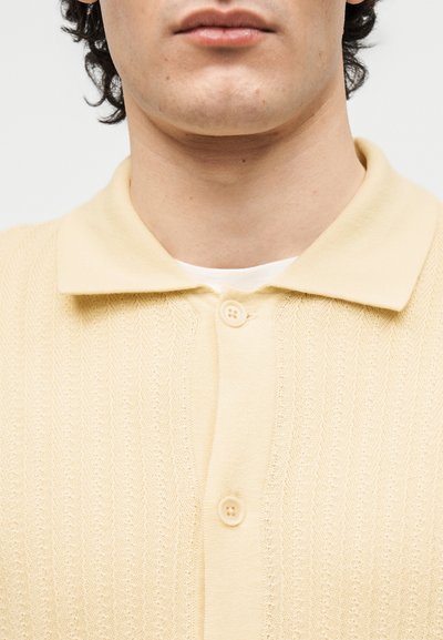 Κοντινό πλάνο ενός ατόμου που φοράει χρωματιστό πλεκτό polo μπλουζάκι σε απαλό κίτρινο χρώμα, κουμπωμένο, πάνω από λευκό εσώρουχο, με ορατό λαιμό και κάτω μέρος προσώπου.