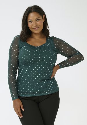 Top de manga larga en color azul aguamarina con lunares blancos, con escote en forma de V y mangas de malla transparente. Combinado con pantalones negros.