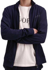 Giacca zip-up blu navy con colletto rialzato, maniche lunghe, tasche frontali, indossata sopra una maglietta bianca con lettering nero in grassetto.
