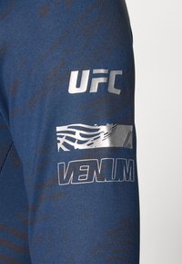 Blå treningsskjorte med en teksturert design, med sølvfargede logoer for UFC og Venum på venstre erme. Stoffet ser glatt og tettsittende ut.