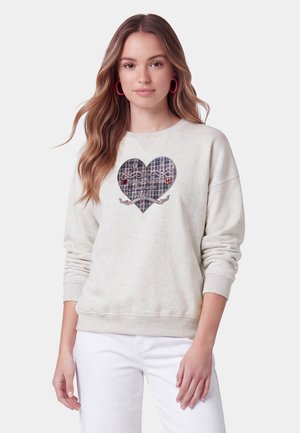 Giovane donna con capelli lunghi indossa una felpa crema con cuore a quadri e testo, abbinata a pantaloni bianchi e orecchini a cerchio rosa, su sfondo semplice.