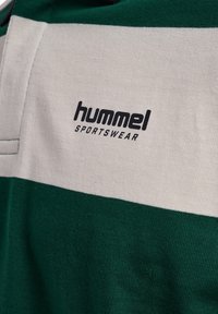 Zielona i biała koszulka sportowa z teksturowanym dzianinowym wykończeniem. Na przodzie znajduje się czarne logo "hummel" oraz napis "SPORTSWEAR".