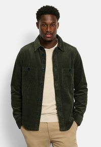 Green corduroy shirt jacket with two chest pockets, featuring dark buttons and a textured fabric. Worn over a light sweater.