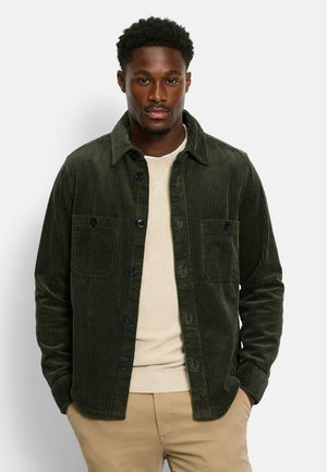 Green corduroy shirt jacket with two chest pockets, featuring dark buttons and a textured fabric. Worn over a light sweater.