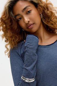 Marineblå og hvid stribet langærmet top med tekstureret stof, med bred halsudskæring og armring på overarmen. Krøllet hår synligt.
