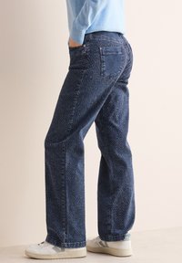 Jeans azul oscuro con un sutil patrón texturizado y corte recto, que cuentan con bolsillos y una cinturilla clásica. Combinados con zapatillas blancas.