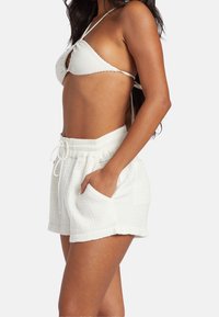 Biały bikini z teksturowanym topem, mającym skrzyżowane ramiączka, oraz lekkie białe szorty z wysokim stanem, wyposażone w elastyczny pas i kieszenie.