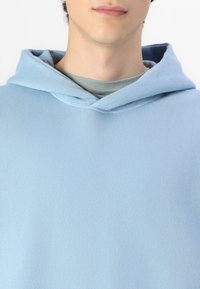 Hanorac din bumbac de culoare albastru deschis, cu un finisaj texturat, prevăzut cu glugă ajustabilă și un aspect stratificat peste o cămașă gri de dedesubt.