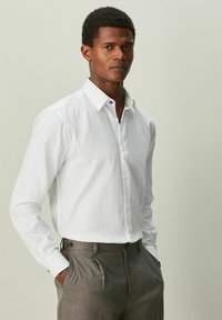 Bela srajca s teksturiranim vzorcem in kontrastnim ovratnikom z vzorcem. Oblečene s sivimi krojenimi hlačami, roke v žepih.