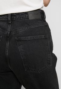 Jeans in denim neri con vita alta e una texture liscia, dotati di tasche posteriori e di un'etichetta in pelle nera con la scritta "WEEKDAY".