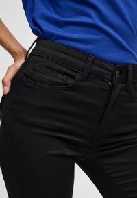 Jeans ajustados negros con una textura suave, que presentan un bolsillo frontal, trabillas para el cinturón y una cremallera. Combinados con una camiseta azul.