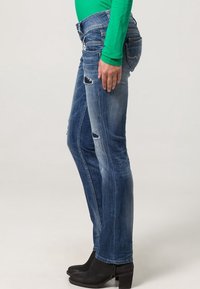 Jeans de mezclilla azul ajustados con detalles desgastados, con una cintura baja y un ajuste ceñido, combinados con botas negras hasta el tobillo.
