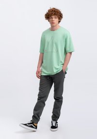 T-shirt oversize verde menta con una grafica a triangolo al centro, abbinato a pantaloni grigi scuri a sigaretta e sneakers nere con dettagli bianchi.