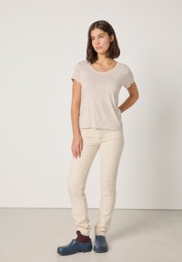 Mladi ženski stoji z eno roko na boku, oblečena v bež majico in hlače, temne nogavice ter modre natikače na enobarvnem ozadju.