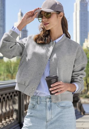 Graue Fleece-Bomberjacke mit gerippten Bündchen, getragen über einem hellblauen Hemd. Hält eine schwarze Kaffeetasse in der Hand, kombiniert mit hellblauen Jeans.