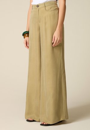 Pantaloni beige a gamba larga indossati con un top bianco, accessoriati con braccialetti verdi e marroni grossi e sandali neri con punta aperta.