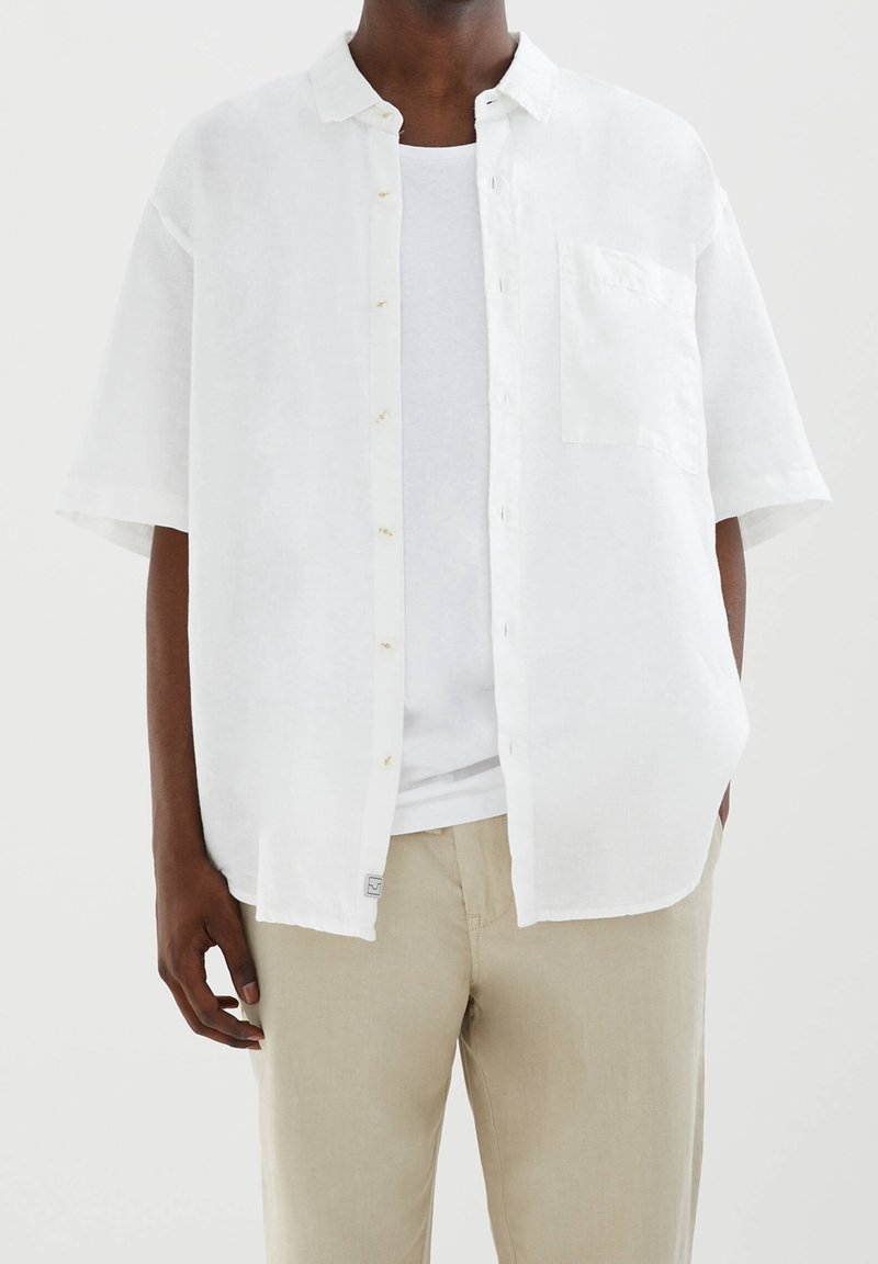 Homme portant une chemise blanche ample à manches courtes boutonnée par-dessus un t-shirt blanc et un pantalon beige, debout avec une main dans la poche.