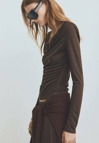 Top maro cu mâneci lungi, cu un decolteu drapat și plii laterale, asortat cu o fustă și un șal legat la talie.