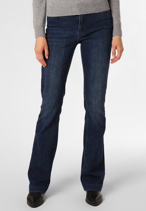 Vrouw draagt donkerblauwe bootcut jeans, grijze trui en zwarte laarzen met spits toelopende neus, staand tegen een effen achtergrond.