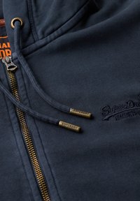Marineblauwe rits-hoodie met een messing rits en metalen koorduiteinden. Kenmerkt zich door een gestructureerde stof en subtiele logo-geborduurde details.
