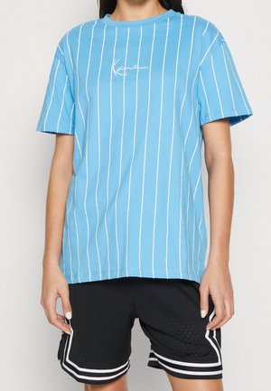 Person iført en blå kortærmet skjorte med hvide lodrette striber og sorte atletikshorts med hvide kanter, stående foran en ensfarvet baggrund.