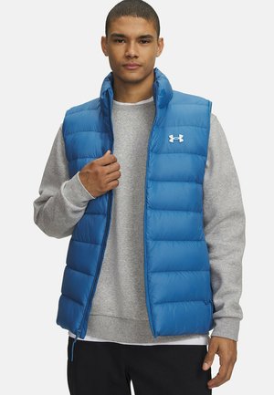 Jeune homme portant une veste matelassée bleue sur un sweat-shirt gris, tenant la fermeture éclair de la veste près du col sur un fond blanc uni.
