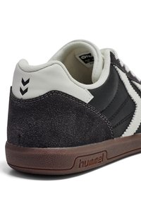 Baskets Hummel en noir et blanc avec talon en suède, semelle marron, et logo distinct à l'arrière ainsi que design chevron sur le côté, vues de profil et de dos.